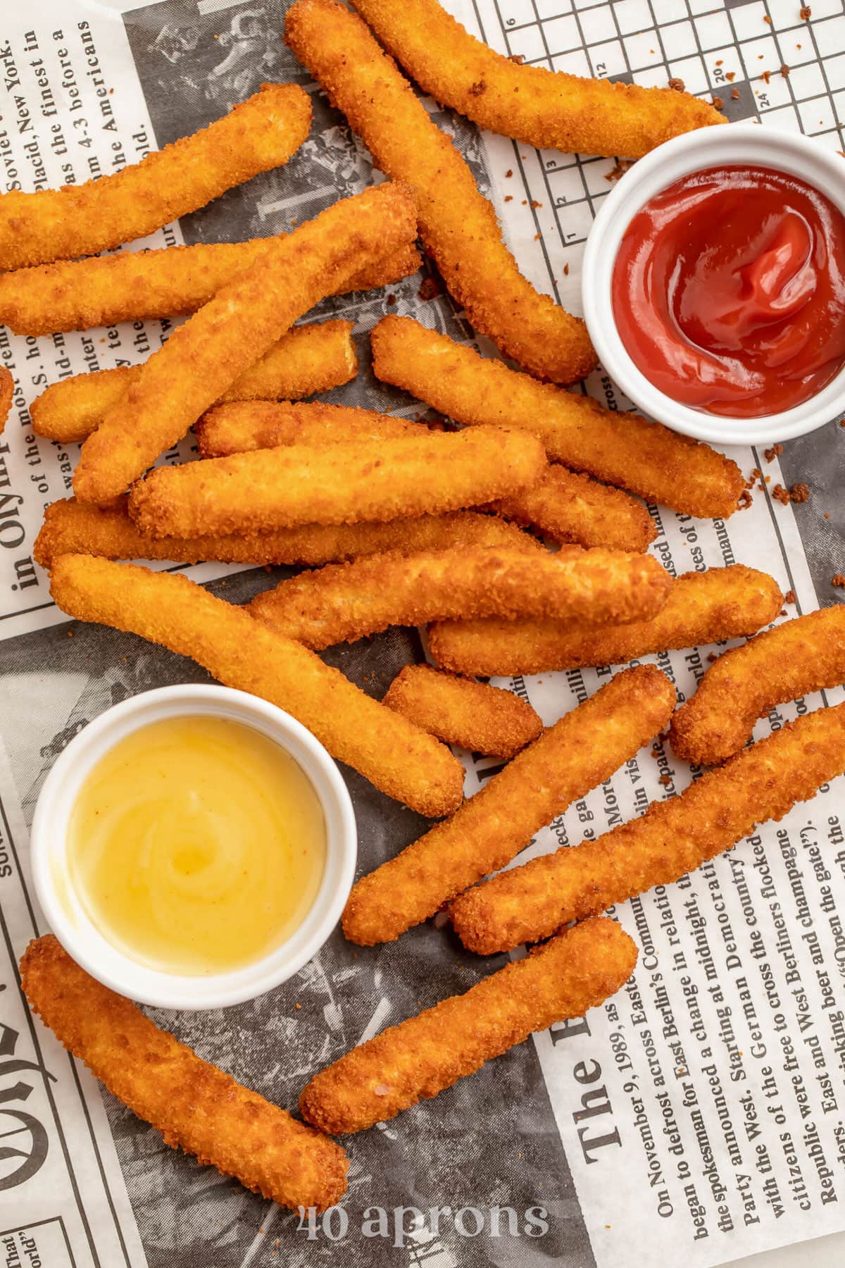 Air fryer cooked chicken fries on a sheet on newspaper next to ramekins of honey mustard and ketchup.