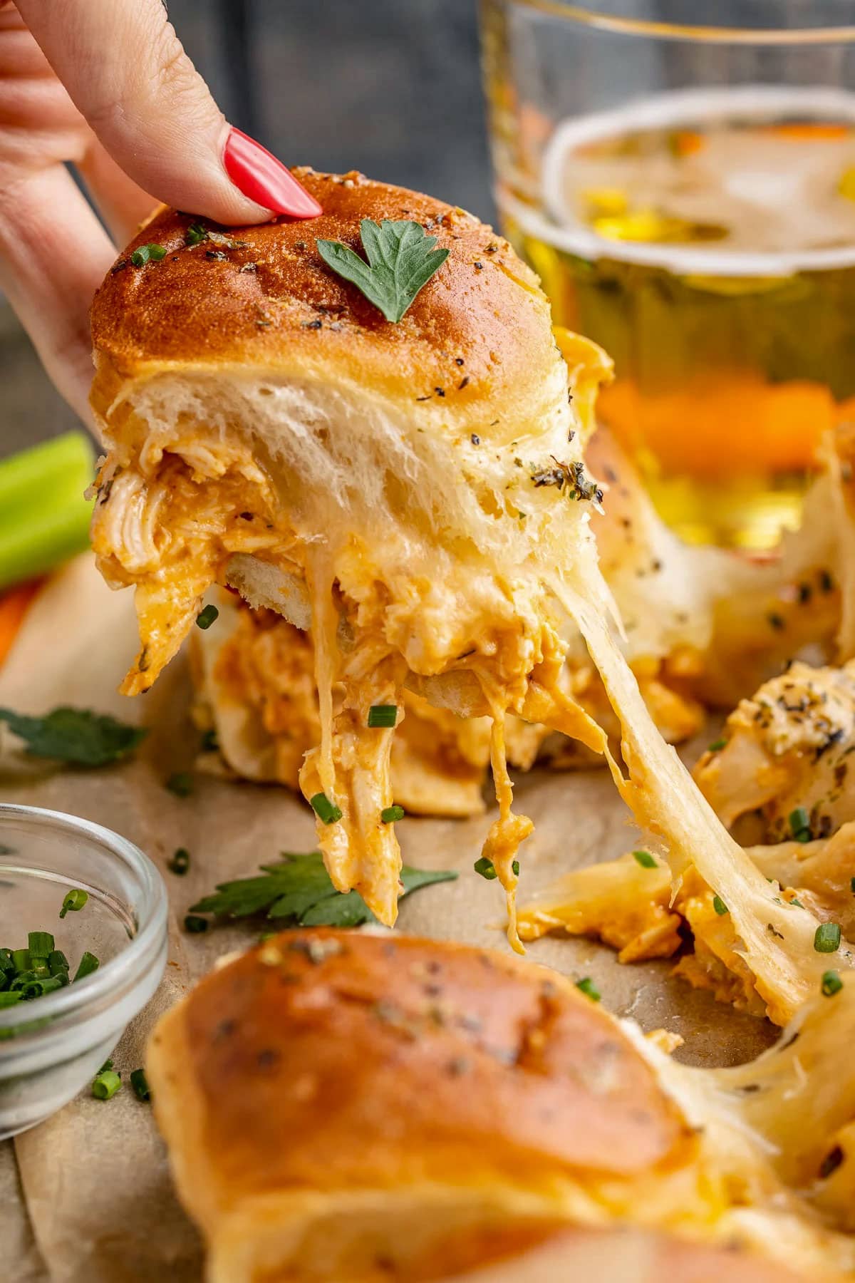 A white woman's hand pulling a buffalo chicken slider away from a sheet pan, with a long pull of melty cheese connecting the sandwiches.