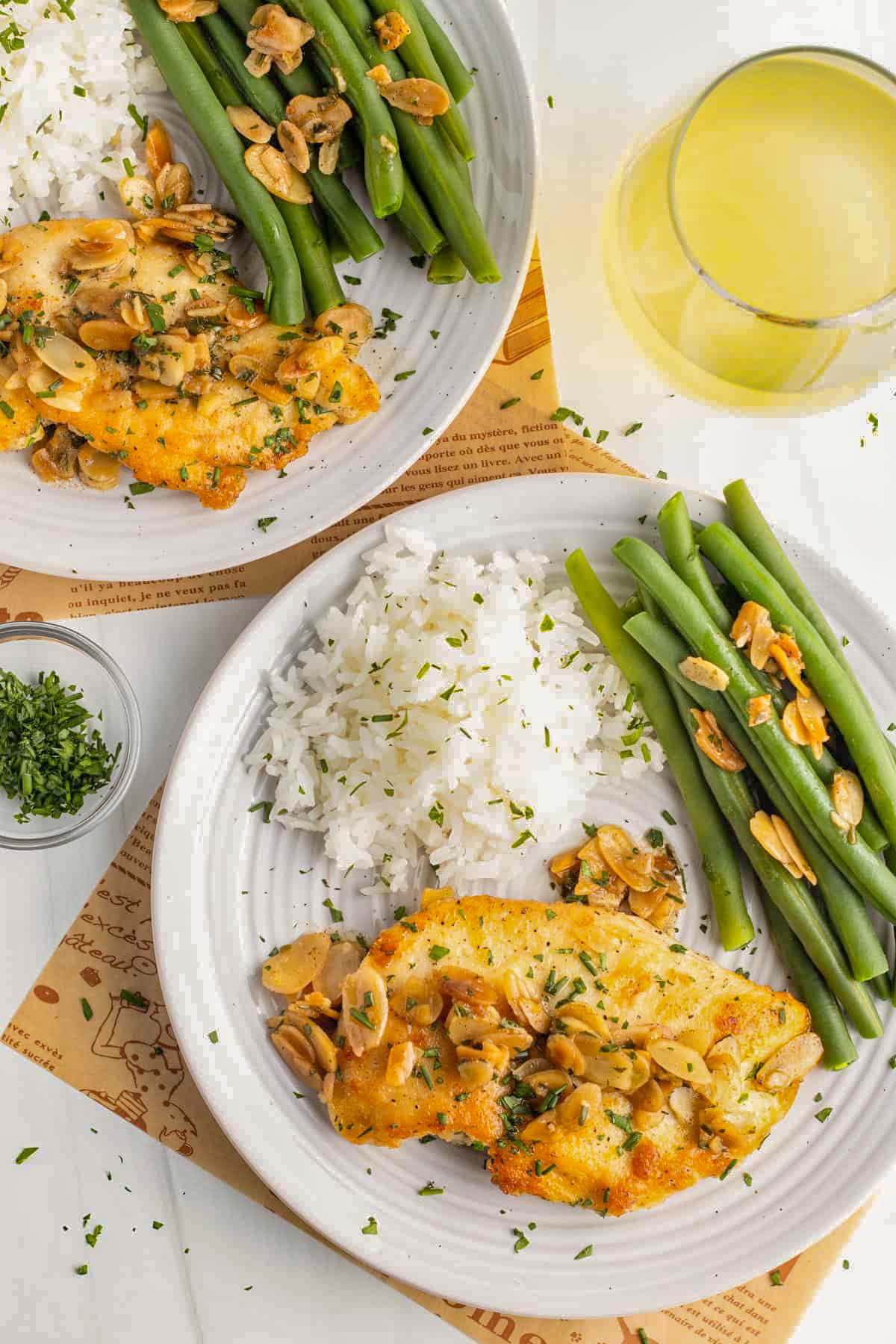 Chicken almondine on a white plate with white rice, green asparagus, almond slivers, next to a glass of white wine.