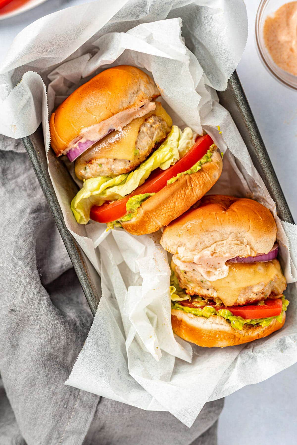 Two chicken burgers, fully dressed, laying on their sides on a baking pan lined with parchment paper.