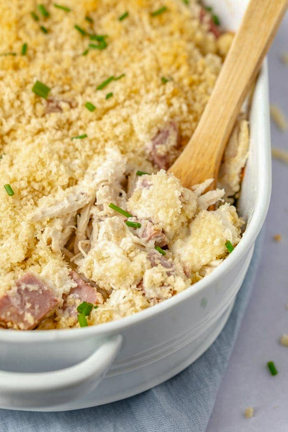 A chicken cordon bleu casserole in a large white oval casserole dish, with a wooden spoon holding a scoop of casserole resting in the dish.