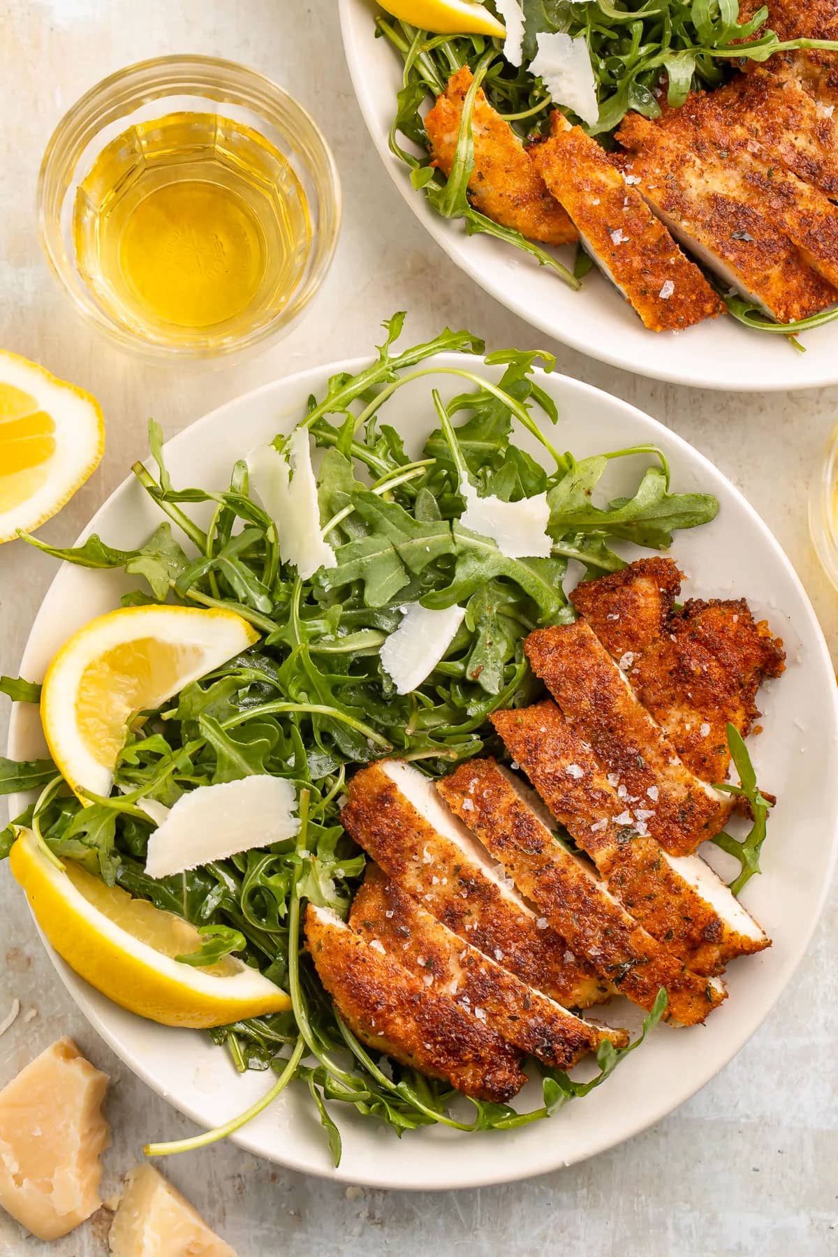 Top-down view of crispy breaded chicken milanese, sliced and plated next to a small green leaf salad with parmesan shavings and lemon wedges, on a table.