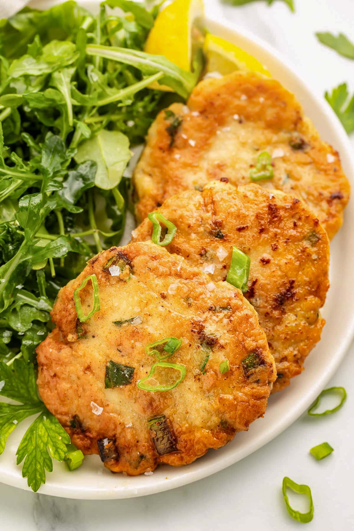 Three round crispy chicken patties on a plate next to a small green leaf salad.