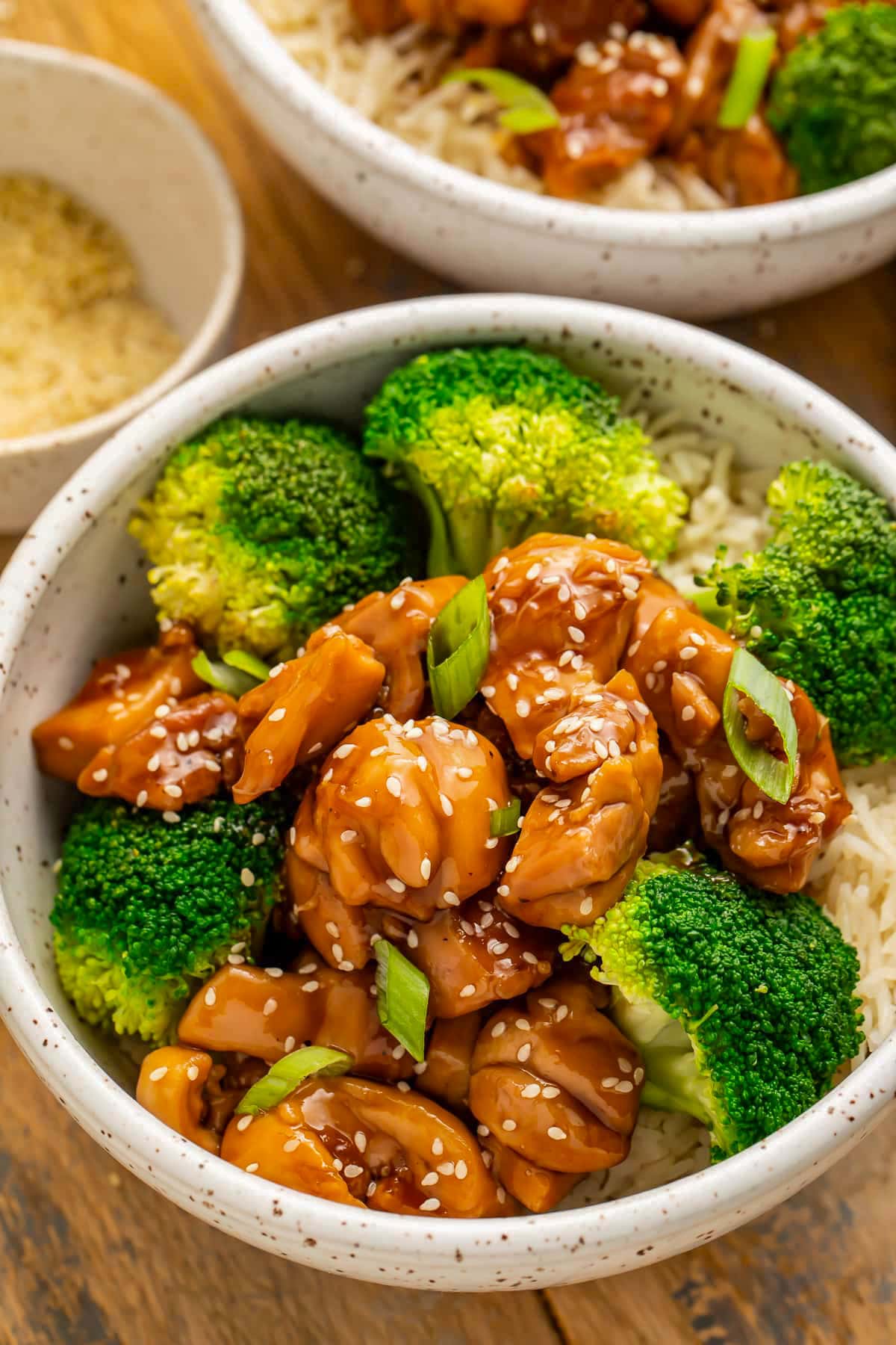 A white bowl holding white rice, bright green steamed broccoli florets, sticky glazed chicken teriyaki, and a sprinkle of white sesame seeds.