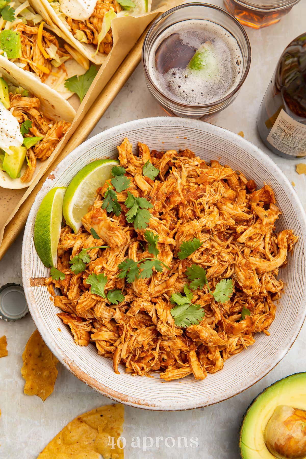 A white bowl holding shredded salsa chicken cooked in an Instant Pot next to a sheet pan of tacos.