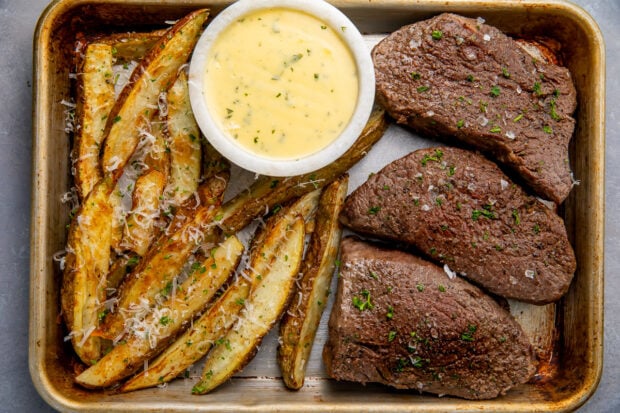 Medium rare steak and thick cut fries with a bowl of bearnaise sauce on a sheet pan