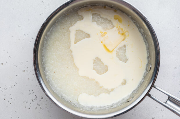 Overhead view of grits, water, butter, and heavy cream in a large silver saucepan.