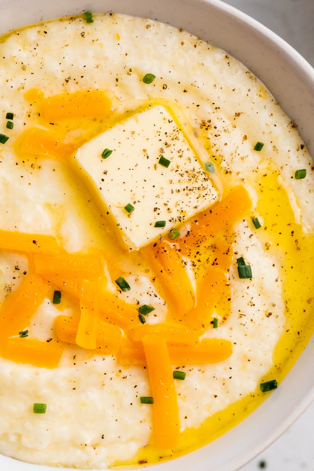 Overhead, close-up view of a bowl of grits with shredded cheese, a pat of butter, and black pepper.