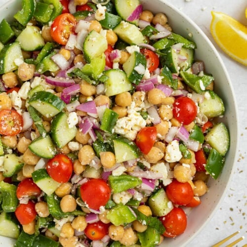 A large white bowl holding a Mediterranean-style chickpea salad with feta, bright red cherry tomatoes, chickpeas, cucumber, onion, and bell pepper.