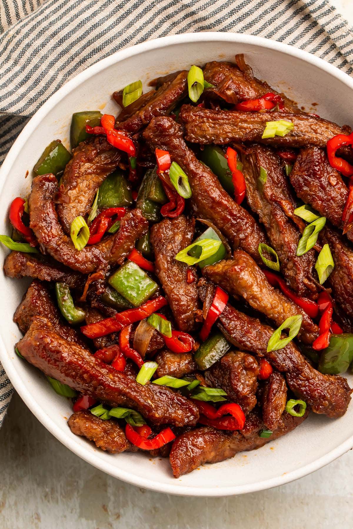 A large white bowl holding strips of hunan beef with vibrant red peppers and green onions.