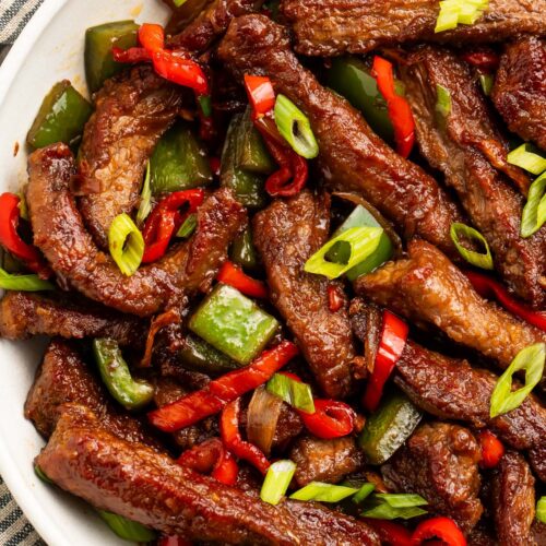 A large white bowl holding strips of hunan beef with vibrant red peppers and green onions.