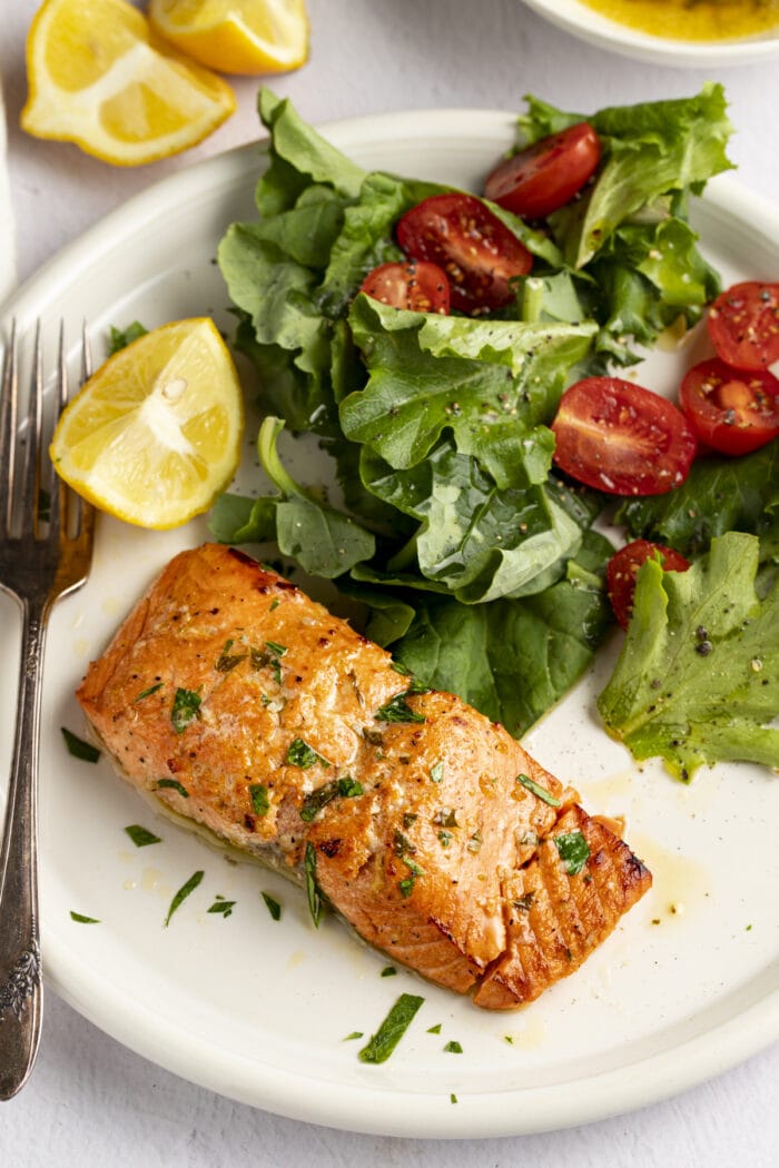 Broiled salmon on a plate next to a small salad and lemon wedges