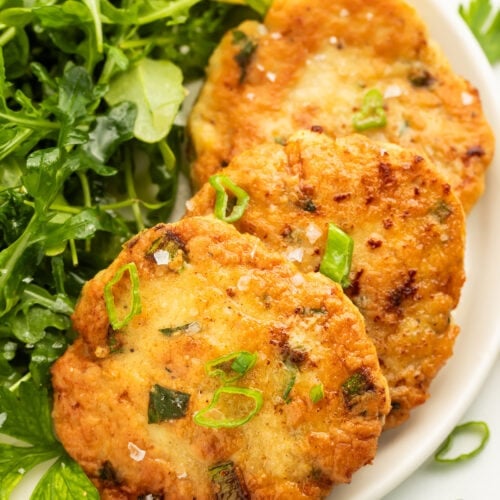 Three pan-fried, golden chicken patties arranged on a white plate with a leafy green side salad.