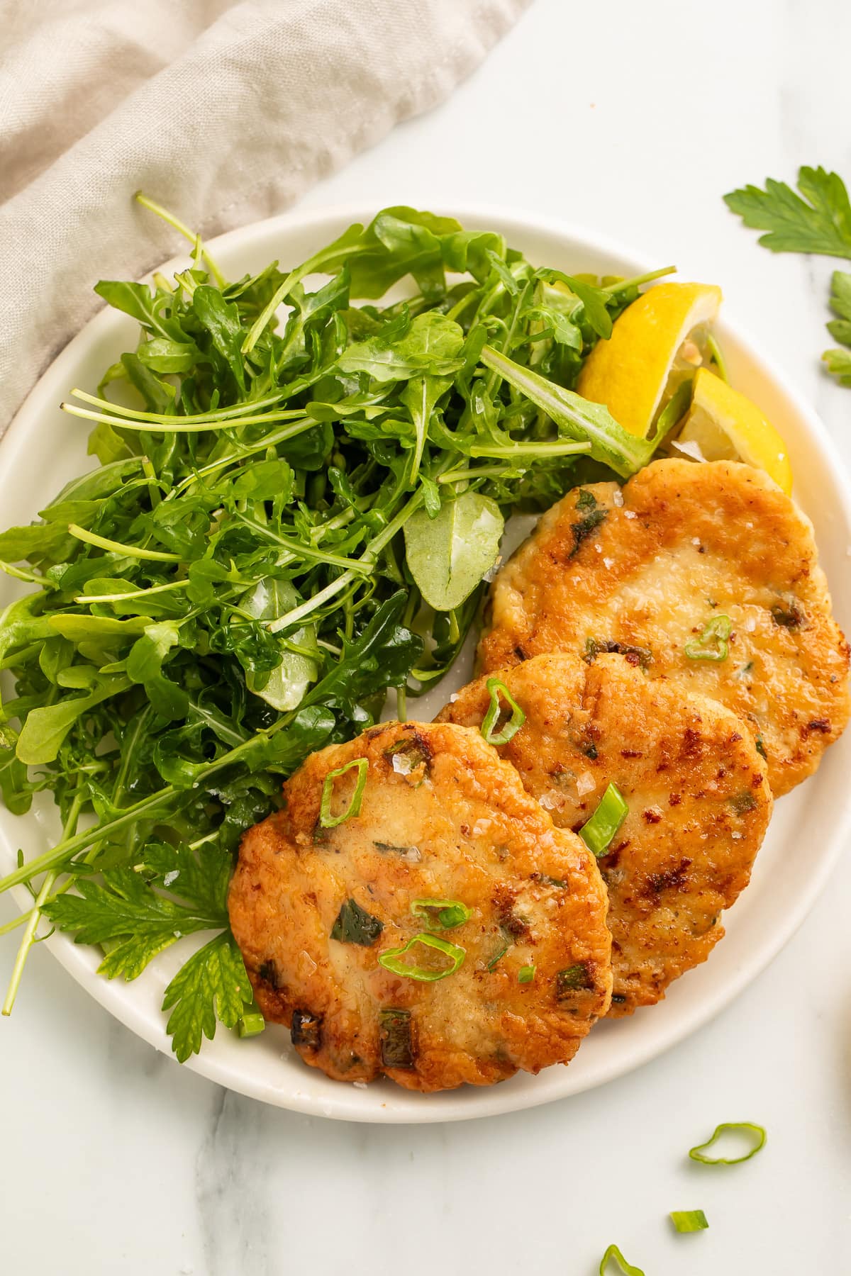 Zoomed out overhead view of three pan-fried, golden chicken patties arranged on a white plate with a leafy green side salad.