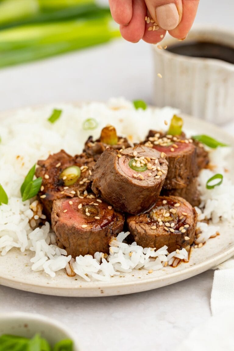 beef negimaki on a plate with rice and sesame seeds being sprinkled over the top