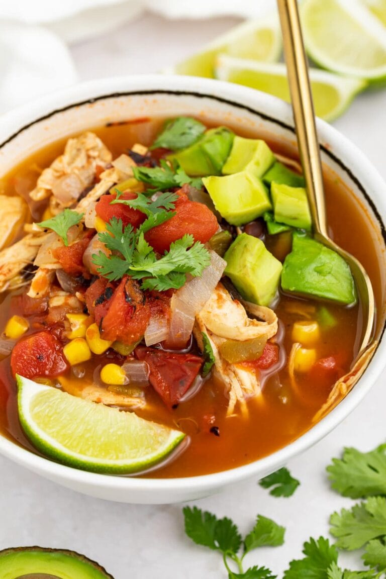 mexican soup in a bowl with a spoon and lime wedges, cilantro, and half an avocado on the side