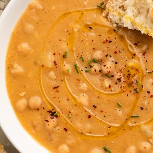 Top-down close-up of a white soup bowl holding creamy, golden chickpea soup with a piece of crusty bread resting in the soup.