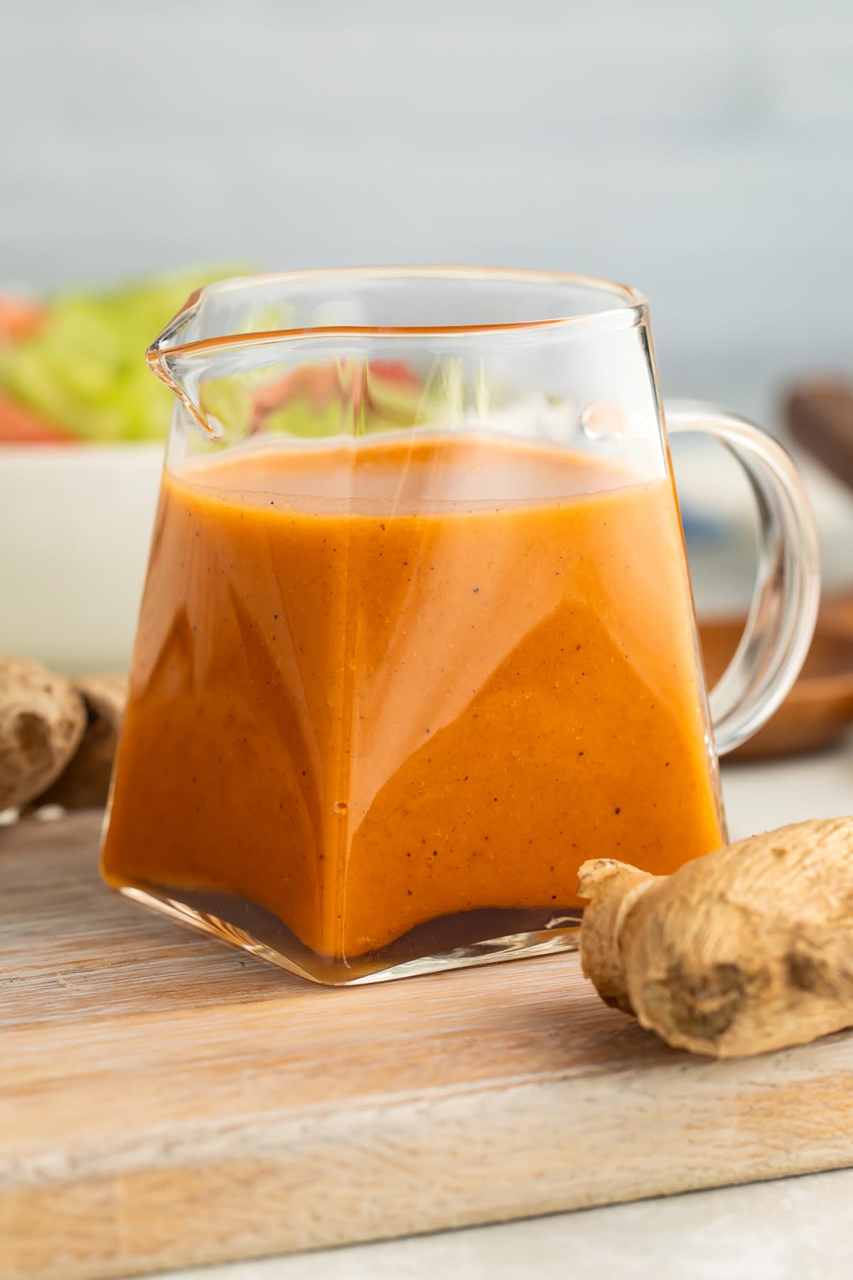 Orange-colored creamy ginger salad dressing in a small clear glass pitcher on a table in front of a bowl of salad.