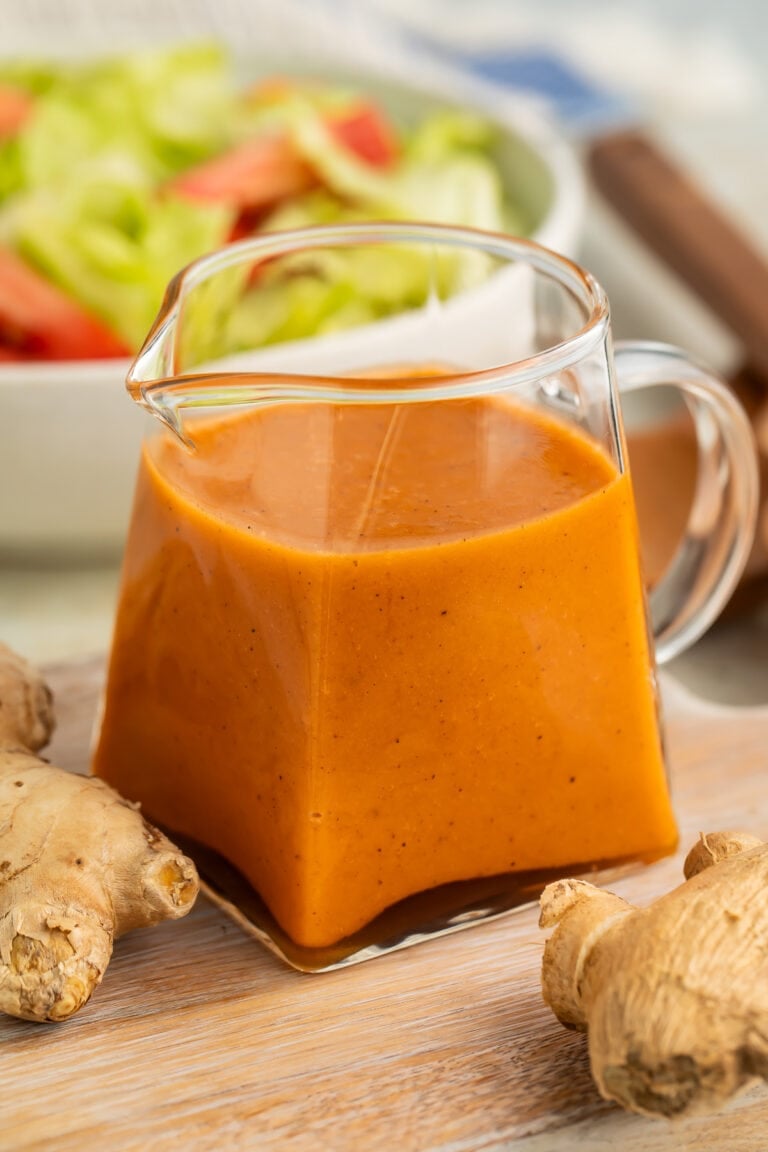 Orange-colored creamy ginger salad dressing in a small clear glass pitcher on a table in front of a bowl of salad.