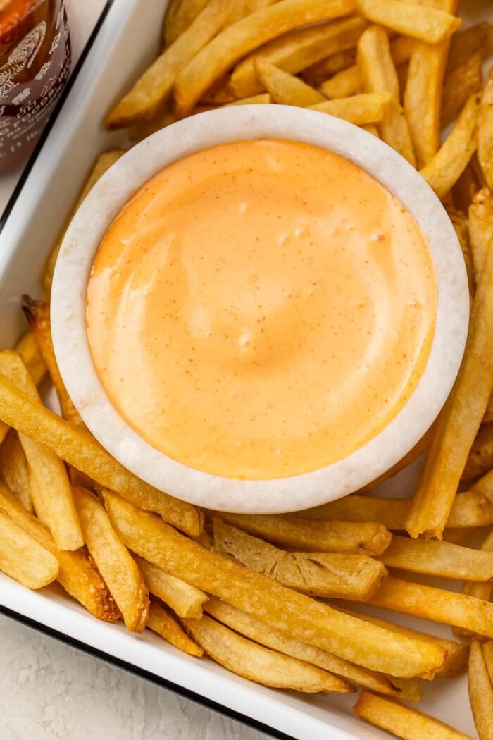 A small bowl of sriracha mayo on a baking sheet surrounded by french fries