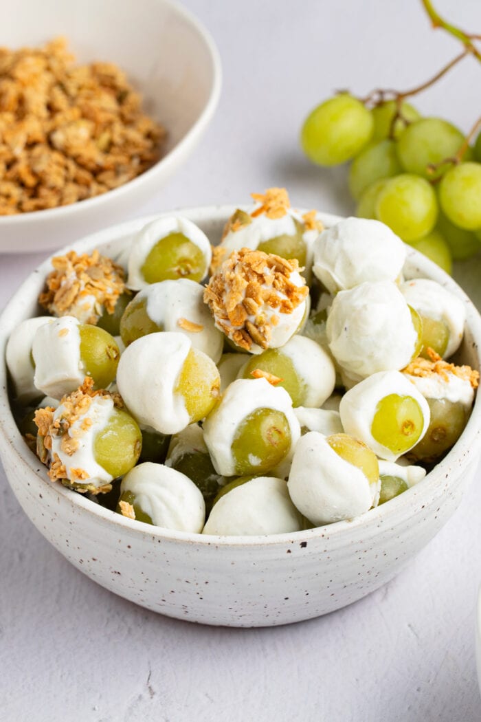 Frozen yogurt-covered grapes in a bowl with grapes and a bowl of granola in the background