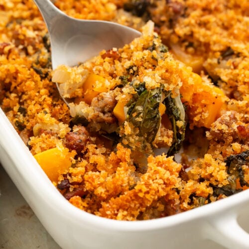 A scoop of butternut squash casserole being lifted out of a casserole dish with a large silver spoon.