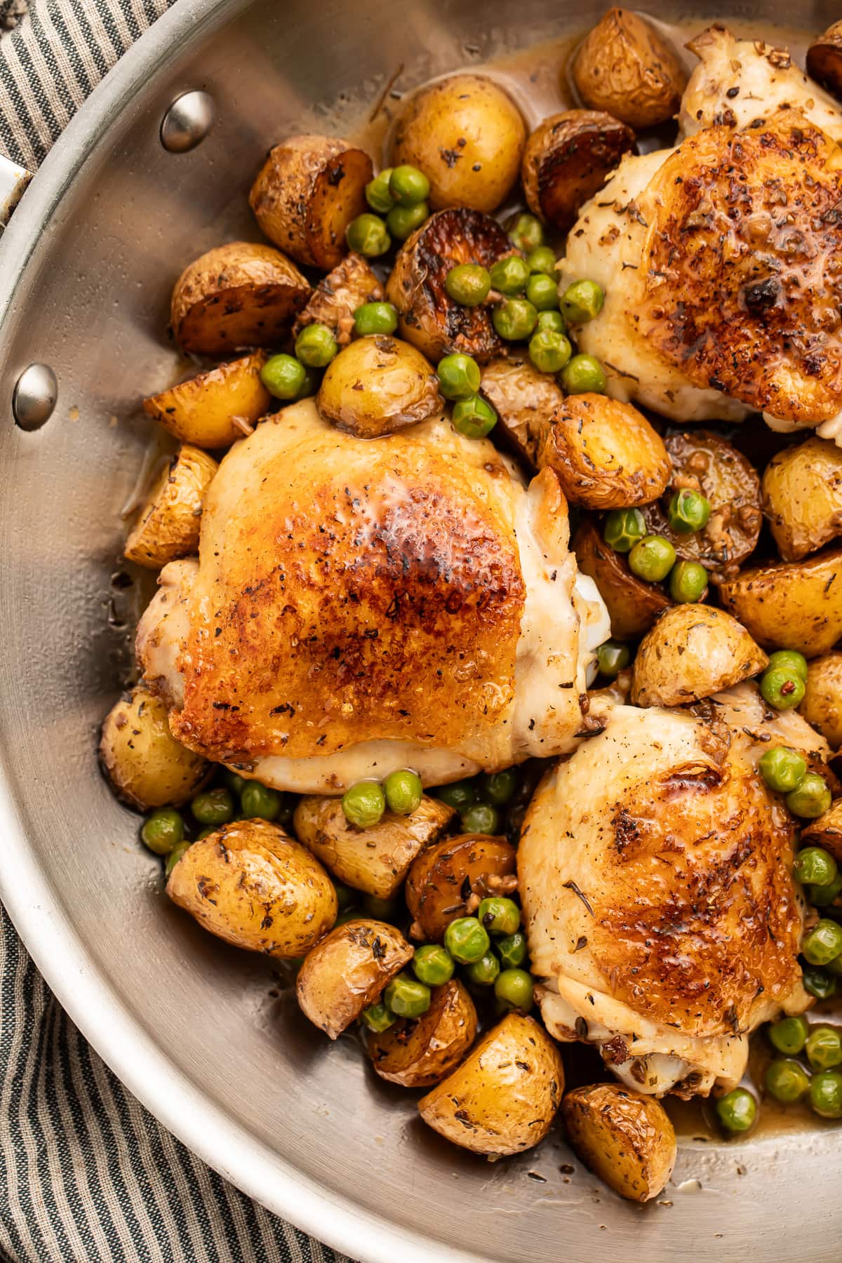 Chicken vesuvio with potatoes and peas in a large silver skillet resting on a striped kitchen towel.