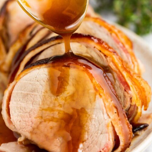 bacon wrapped pork tenderloin on a plate with brown sugar glaze being spooned over the top