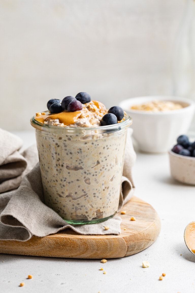 A glass of peanut butter overnight oats topped with peanut butter and blueberries on a wooden serving board.