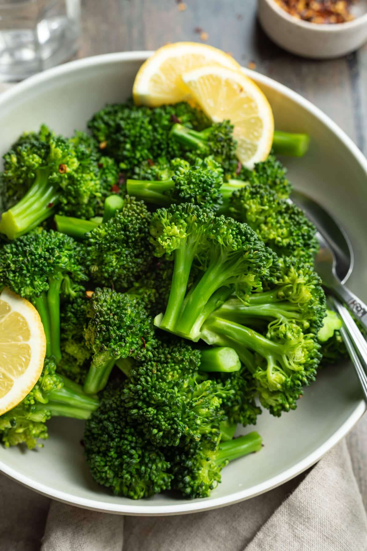 Overhead view of a bowl of steamed broccoli florets with leon wedges on a wooden tabletop.