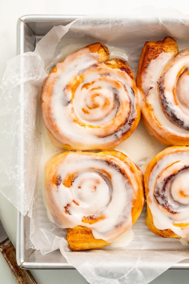 4 air fryer cinnamon rolls topped with melty cinnamon roll icing resting in a pan lined with parchment paper.