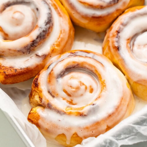 4 air fryer cinnamon rolls topped with melty cinnamon roll icing resting in a pan lined with parchment paper.