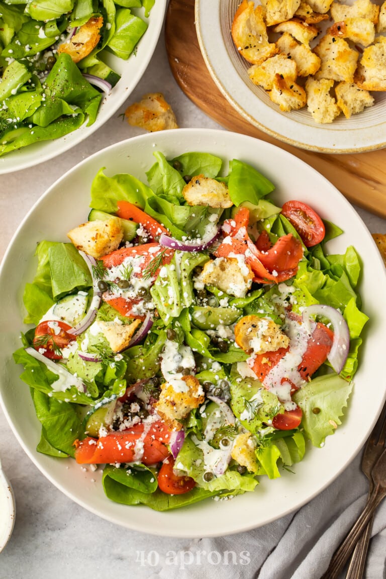 Overhead view of a smoked salmon bagel salad in a white bowl with croutons in another bowl.