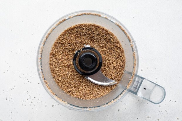 Overhead view of finely chopped pecans in a food processor bowl on a white background.