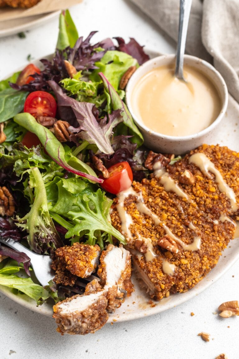 Close-up, angled view of pecan crusted chicken with honey mustard sauce and small side salad on a white plate.