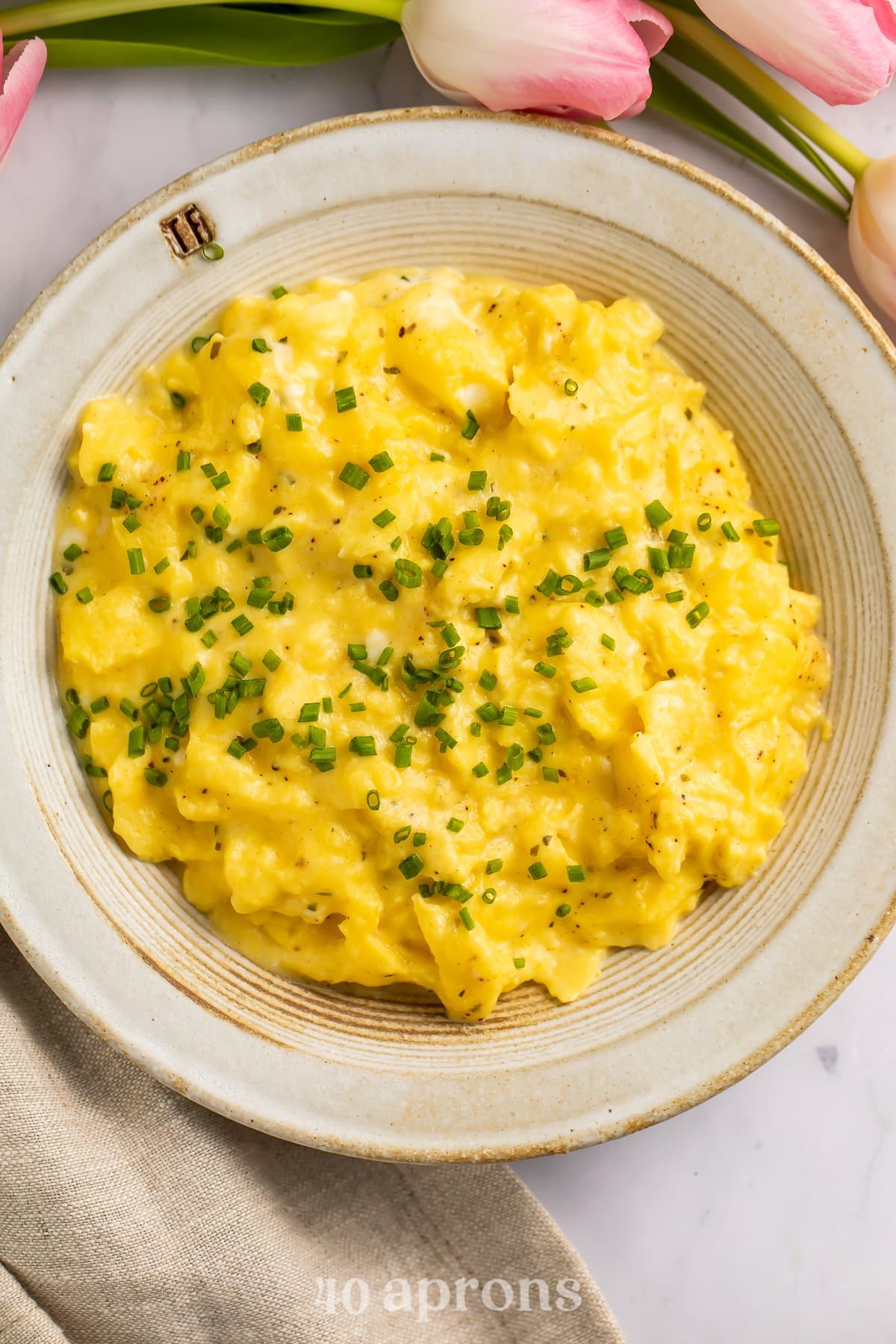 Overhead view of a bowl of boursin scrambled eggs garnished with chives on a neutral countertop.