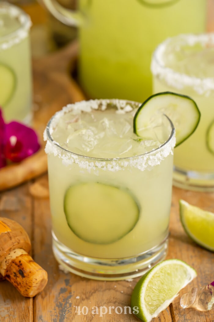 3/4-angle overhead view of a rimmed glass with cucumber margarita and lime wedges.