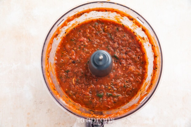 Overhead view of red roasted salsa in a food processor bowl on a light neutral countertop.