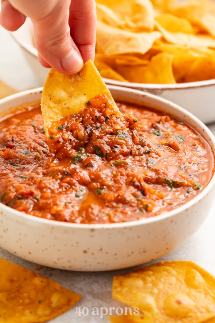 A tortilla chip being dipped into a white bowl containing red roasted salsa.