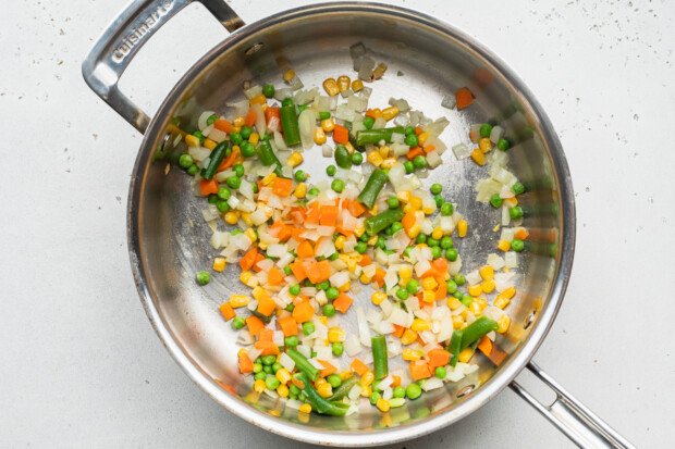 Overhead view of a large skillet with avocado oil, diced onion, and frozen mixed vegetables.