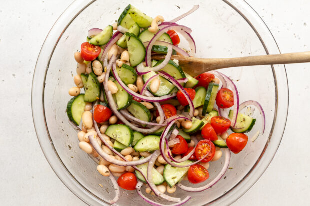 Overhead view of white bean salad ingredients in a large glass mixing bowl with a large wooden spoon.