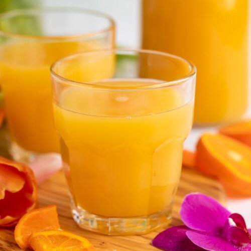 Side view of a glass of bright orange POG juice with a carafe of POG Juice and another glass in the background.