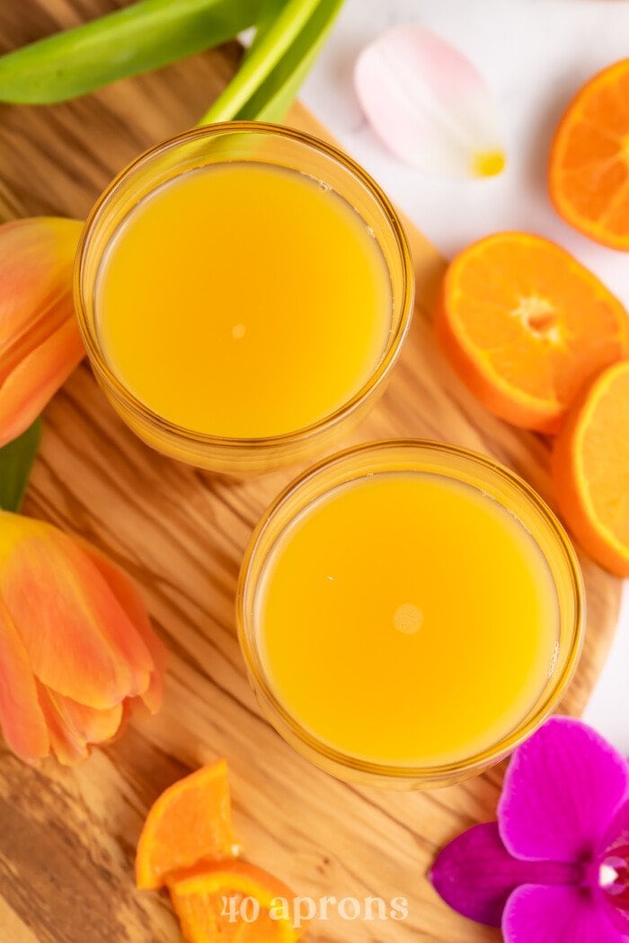 Overhead view of two glasses of POG juice on a wooden serving tray with bright pink tropical flowers.