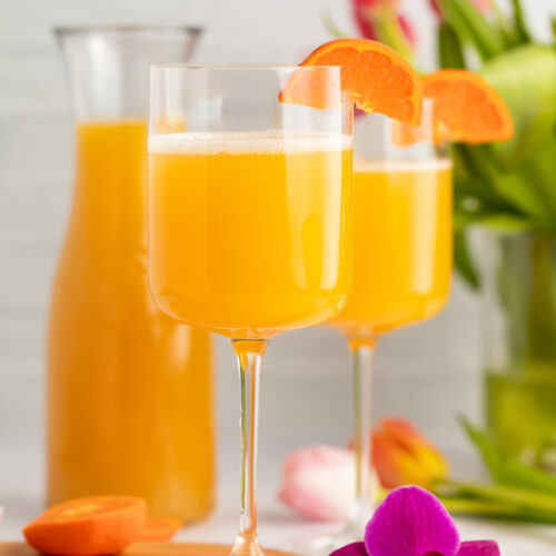 Side view of two wine glasses holding POG juice mimosas with a carafe of pog juice in the background.