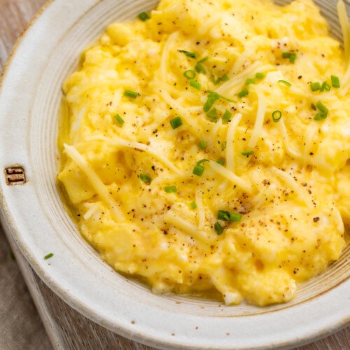 Close-up, overhead view of a bowl of truffled scrambled eggs garnished with chives on a wooden serving board.