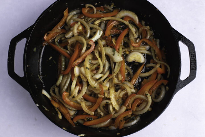 Overhead view of sauteed peppers, onions, and fresh herbs in a large cast iron skillet on a white table.