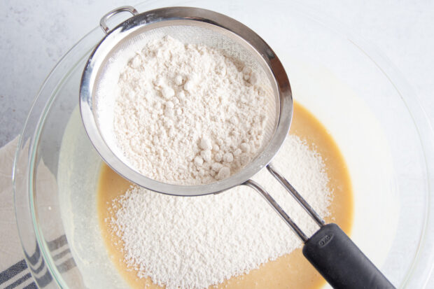 Dry ingredients for gluten free blueberry muffins in sifter over wet ingredients in large glass mixing bowl.