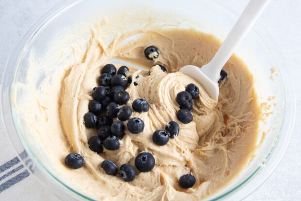 Fresh blueberries in gluten free blueberry muffin batter in large glass mixing bowl with spatula.