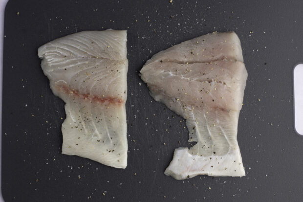 Barramundi fillets seasoned with salt and pepper on a black cutting board.