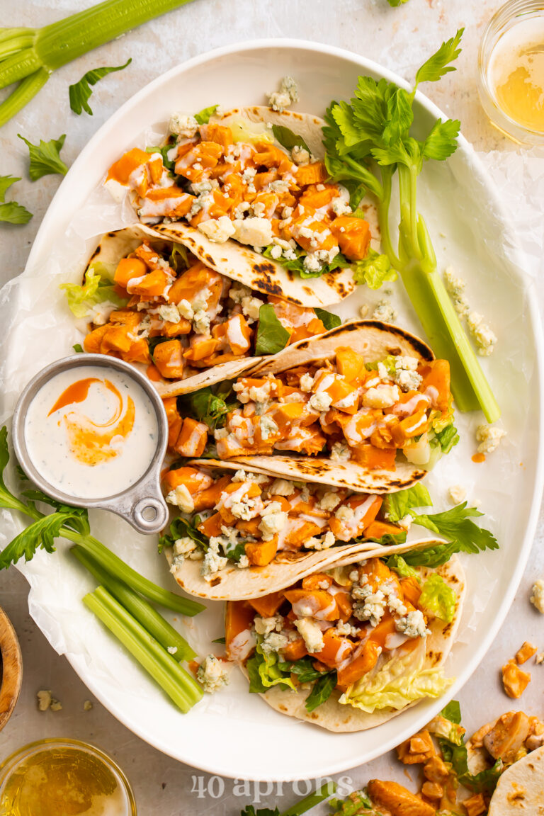 Overhead view of 5 buffalo chicken tacos lined vertically on an oval platter with celery sticks and ranch.